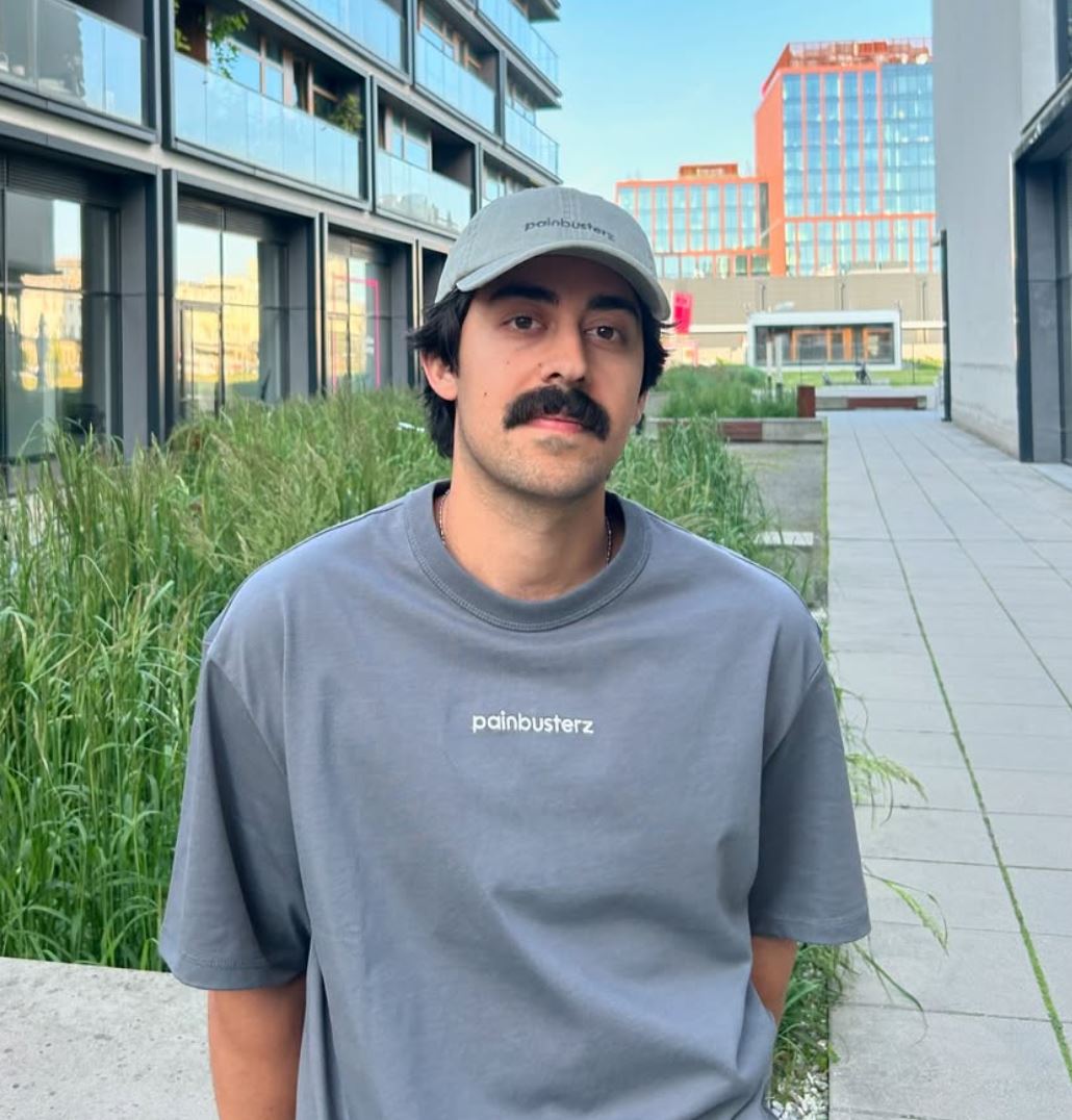 Mory Naimi wearing a grey Painbusterz T-shirt and a grey-green limited edition Painbusterz cap, Summer 2025.