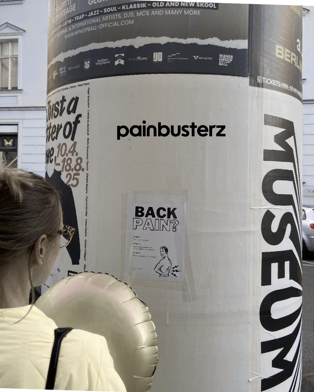 Artistic black-and-white photo of a person stopping to view a small Painbusterz poster about back pain among other city posters.